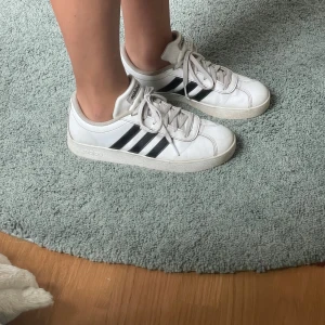 Vita Adidas sneakers med svarta ränder - Klassiska vita Adidas sneakers med de ikoniska tre svarta ränderna på sidorna. Skorna har snörning, rund tå och platt sula. Perfekta för en sportig och avslappnad stil. Tillverkade i syntetmaterial och skinnliknande yta.