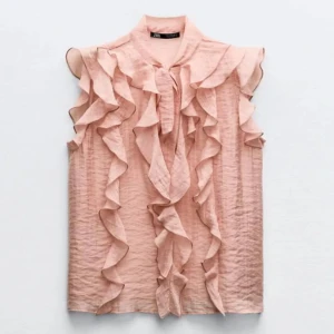 Ljusrosa volangblus med rosett Zara - Supersöt ljusrosa blus från Zara med volanger längs axlar och framtill. Blusen är ärmlös och har en snygg rosettdetalj vid halsen som man kan knyta . Storlek xs🫶🏻 en liten defekt finns se sista bilden, men är på insidan på en av volangerna så man märker det inte om man inte ”vänder” på volangen och kollar väldigt noga❤️