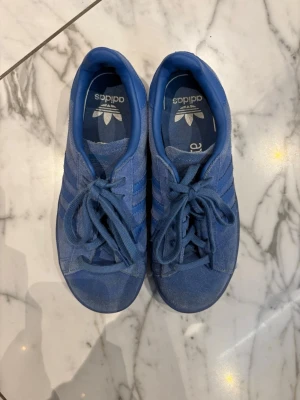 Blå adidas Gazelle sneakers mocka - Säljer ett par blå adidas Gazelle sneakers i mocka med klassiska tre ränder på sidan. Skorna har snörning och platt sula, samt adidas-logga på plösen och hälen. Perfekta för dig som gillar retrostil och vill ha något bekvämt och snyggt.