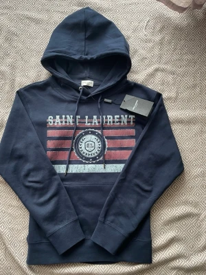 Mörkblå Saint Laurent hoodie - Snygg mörkblå hoodie från Saint Laurent med tryck i vitt och rött framtill. Klassisk huva med dragsko och ribbade muddar. Märkets logga syns tydligt på bröstet och etiketten sitter kvar. Perfekt för dig som vill ha en stilren och trendig look.