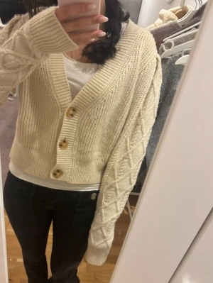 Beige stickad kofta från Bik Bok - Mysig beige stickad kofta från Bik Bok med grov ribbstickning och dekorativa knappar framtill. Koftan har ett croppat snitt, v-ringning och mönstrade ärmar.