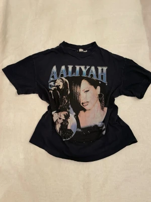 Mörkblå/nästan svart Aaliyah t-shirt med tryck - Cool mörkblå/svart t-shirt med stort Aaliyah-tryck på framsidan i blått och vitt. Klassisk rund hals och korta ärmar. Perfekt för dig som gillar streetstyle och vill ha en unik t-shirt med ikonisk artist.