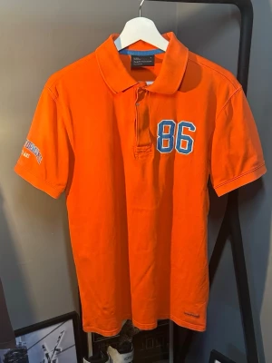 Orange pikétröja med tryck från PeakPerformance - Snygg orange pikétröja från PeakPerformance med blåvitt '86'-tryck på bröstet och broderad text på ärmen. Klassisk krage och knäppning framtill. Tillverkad i bomull för en bekväm och luftig känsla. Perfekt för en sportig och avslappnad stil.