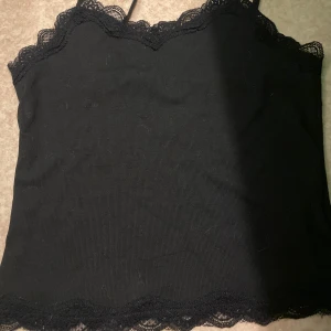 Svart spetskantad linnetopp - Svart linnetopp med smala axelband och dekorativ spets längs både halsringning och nederkant. Perfekt att styla med jeans eller kjol för en trendig look.