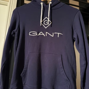 Mörkblå hoodie från GANT - Mörkblå hoodie från GANT med vit broderad logga på bröstet och vita dragsnören i huvan. Klassisk känguruficka framtill och mjukt material som känns skönt mot huden. 