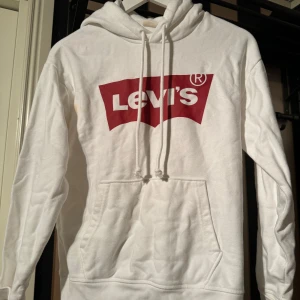 Vit hoodie från Levi's med logga - Vit hoodie från Levi's med den klassiska röda loggan framtill. Tröjan har huva med dragsko och en stor magficka. 