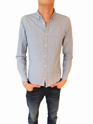 Ljusblå skjorta med knappar - Klassisk ljusblå skjorta med lång ärm och vita knappar. Skjortan har en smal passform och button-down krage, perfekt för en clean och stilren look. Tillverkad i bomull för en bekväm känsla hela dagen.