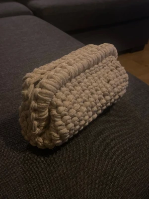 Beige stickad kuvertväska - Handgjord beige kuvertväska i grovt stickat material. Väskan har en rektangulär form och öppnas upptill. Perfekt accessoar för dig som gillar unika och trendiga detaljer.
