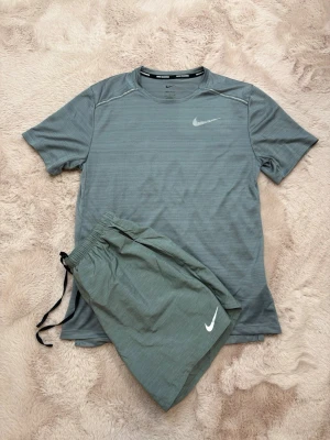 Nike Shortsset - Nike träningsset med grå t-shirt och matchande shorts. T-shirten har rund hals, kort ärm och Dri-FIT-material som andas. Shorts med resår och dragsko i midjan. Båda plaggen har Nike-logga framtill. Perfekt för löpning eller gym. T-Shirt storlek: S Shorts storlek: M