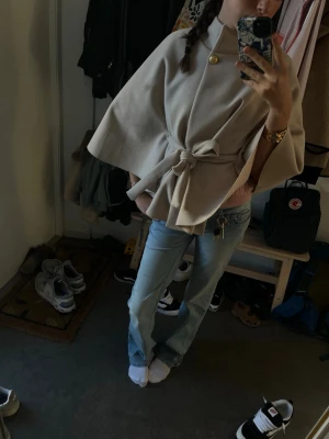 Beige kappa med bälte och vida ärmar - Stilren beige kappa med knytbälte i midjan och vida, trekvartsärmar. Kappan har en elegant och enkel design som ger en snygg siluett. Perfekt för dig som vill ha en trendig och tidlös look.