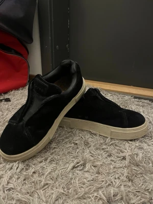 Svarta sneakers från Etys - Stilrena svarta sneakers från etys med beige sula och lila detalj baktill. Ovandelen är i mocka och skon har en låg profil utan snörning. Perfekta för en clean och enkel look.