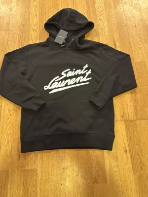 Saint Laurent hoodie - Svart hoodie från Saint Laurent med vit logga i snyggt tryck på bröstet. Storlek S. HELT NY!! 