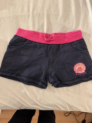 Low waist Mörkblå shorts Abercrombie & Fitch - Mörkblå shorts från Abercrombie & Fitch med bred rosa midjeresår och snörning. Shortsen har två fickor fram och ett broderat märke i rosa och gult på vänster ben. Perfekta för sommardagar och avslappnade tillfällen.