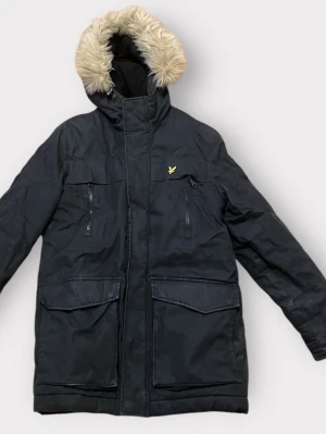 Svart parkasjacka med fuskpäls - Svart parkasjacka från Lyle & Scott med huva och fuskpäls. Jackan har dragkedja, stora fickor framtill och en gul logga på bröstet. Perfekt för kalla dagar med sin långa passform och varma material.