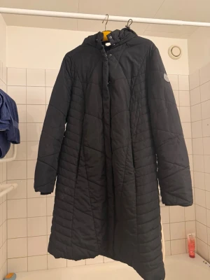 Svart quiltad kappa med huva - Lång svart quiltad kappa från Outdoor med huva och dragkedja framtill. Kappan har ett stilrent mönster och en patch på ärmen. Perfekt för kyliga dagar och enkel att matcha med olika outfits.