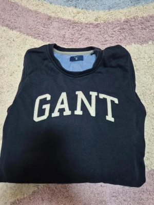 sweatshirt GANT - Säljer en marinblå sweatshirt från GANT med stor vit logotyp broderad på bröstet. Tröjan har rund halsringning och klassisk passform. Perfekt för en avslappnad och stilren look.