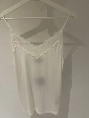 Vit spetslinnetopp från Object storlek S/M - Säljer en vit linnetopp från Object med tunna axelband och vacker spetsdetalj längs v-ringningen. Toppen har en mjuk och lätt känsla, perfekt för lager på lager eller sommarens varma dagar.