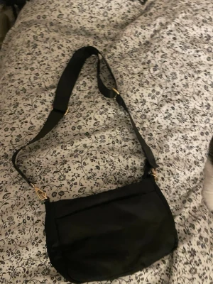 Svart axelväska med bred rem - En stilren svart axelväska med bred justerbar rem och guldfärgade metalldetaljer. Väskan har en enkel, mjuk form och en praktisk ytterficka framtill. Perfekt för dig som vill ha en clean och funktionell look.