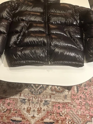 Svart glansig dunjacka med dragkedja - Säljer en svart, glansig dunjacka med quiltad design och hel dragkedja framtill. Jackan har en puffig form och långa ärmar, perfekt för kalla dagar. Snygg och enkel att matcha till olika outfits.