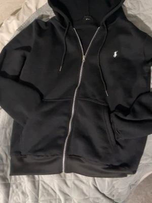 Svart hoodie med dragkedja från Polo Ralph Lauren - Svart hoodie med dragkedja från Polo Ralph Lauren. Klassisk design med vit broderad logga på bröstet, huva med dragsko och två fickor framtill. Tillverkad i mjukt material som är skönt att bära. Perfekt för en avslappnad och stilren look.