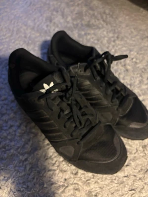 Svarta Adidas ZX 750 sneakers - Snygga svarta Adidas ZX 750 sneakers med klassiska tre ränder på sidan och logga på plösen. Skorna har snörning och är gjorda i en mix av skinn och syntetmaterial med mesh-detaljer för extra komfort. Perfekta för en sportig och stilren look.