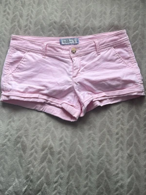 Ljusrosa shorts från Abercrombie - Snygga ljusrosa shorts från Abercrombie & Fitch med klassisk passform och broderad logga på bakfickan. Shortsen har fickor fram och bak, bälteshällor och stängs med knapp och dragkedja. Low waist shorts. Perfekta för varma dagar. Storlek står ej men upplevs som S/M.