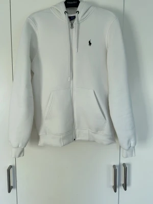 Vit hoodie från Polo Ralph Lauren - Vit Ralph Lauren hoodie. Klassisk design med huva och dragsnören, två stora fickor framtill och den ikoniska svarta loggan broderad på bröstet. Tillverkad i mjukt bomullsmaterial för en skön och avslappnad stil.