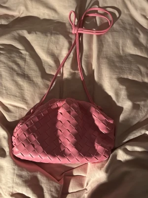 Clutch bag i rosa  - Säljer en superfin rosa handväska med flätad design och lång axelrem som kan knytas. Väskan har en mjuk, rundad form och är tillverkad i syntetmaterial. Perfekt för dig som vill ha en trendig accessoar med det lilla extra.