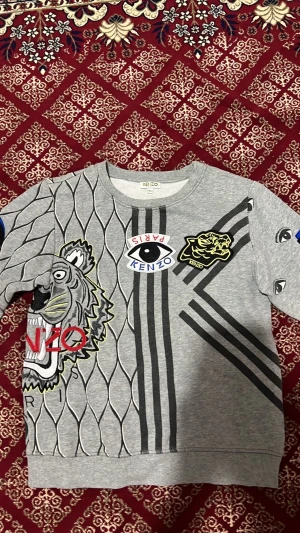 Grå sweatshirt med tryck från Kenzo - Cool grå sweatshirt från Kenzo med stora grafiska tryck, tigrar, ögon och textdetaljer i svart, rött, blått och gult. Tröjan har rund hals, ribbade muddar och unikt mönster både fram och bak. Perfekt för dig som gillar statement-plagg och streetwear.