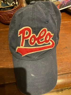 Mörkblå Polo Ralph Lauren keps - Snygg mörkblå keps från Polo Ralph Lauren med stor röd och vit 'Polo'-logga framtill och klassisk röd broderad polospelare på sidan. Kepsen har böjd skärm och justerbar passform. Tillverkad i bomullstyg för en avslappnad och sportig look.