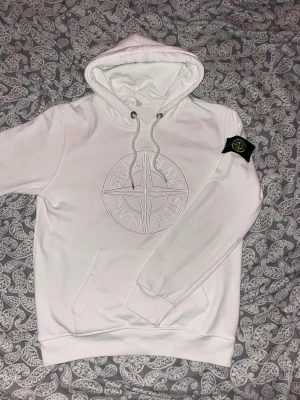 Stone island hoodie - Vit - strl S - nytt skick - Exklusiv vit hoodie från Stone Island i storlek S. Plagget är i nyskick, mycket sparsamt använt och helt utan fläckar, hål eller slitage. Klassisk och eftertraktad modell med den ikoniska kompassloggan diskret broderad på bröstet samt original Stone Island-patch på ärmen.  Tillverkad i högkvalitativ bomull med bekväm passform, huva med dragsko och praktisk magficka. Ett tidlöst plagg som passar både vardag och en mer uppklädd streetwear-stil.