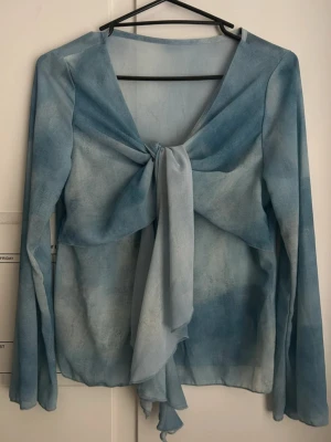 Topp i chiffon - Dazy - Topp i chiffon med volang och knytdetalj Blusen har en v-ringning. Den här blusen är gjord av chiffon och har en relaxed fit och tunt material. Den har en trumpetärm och en knytbar stängning på framsidan med en kråsdetalj.  Storlek: S Färg: Ljusblå  Märke: Dazy Skick: Mycket bra – sparsamt använd, inga fläckar eller skador.