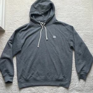 Grå hoodie från Acne Studios - Säljer en stilren grå hoodie från Acne Studios med vit dragsko och diskret logopatch på bröstet. Tröjan är i mjuk bomull och har klassisk huva samt ribbade muddar. Perfekt för en avslappnad look. Taggad som xs men passar som M. 