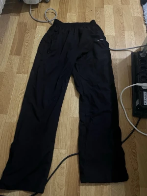 Svarta mjukisbyxor från Rich Studios - Svarta mjukisbyxor från Rich Studios med avslappnad passform och resår i midjan. Byxorna har dragkedjeförsedda fickor och diskret logga på höger sida. Perfekta för chill dagar eller när du vill ha det bekvämt. Elr en skön vintage outfit