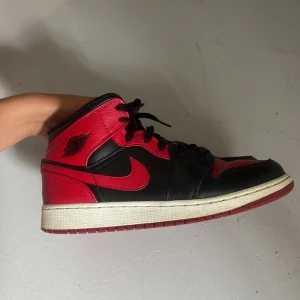Nike Air Jordan 1 svart/röd strl 39 - Säljer ett par Nike Air Jordan 1 mid banned i klassisk svart och röd färg. Skorna har snörning, rund tå och platt sula. Ovandelen är i skinn med ikoniska swoosh-loggan på sidan och Air Jordan-vingen vid ankeln. Perfekt för dig som gillar streetstyle och sneakers med attityd. Skorna är äkta och köpta för ett tag sen men kommer ej till användning längre