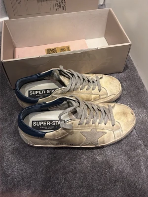 Golden Goose Superstar sneakers beige - Golden Goose Superstar sneakers i beige med grå snörning, ikonisk stjärndetalj på sidan och mörkblå häl. Ovandelen är i skinn med en vintage-look och platt sula. Klassisk låg modell med rund tå och tydlig branding på insidan.ett par rätt slitna skor men går säkert att få tillbaka i liv lite mer köpta för 3999 säker for 799 allt på bilderna ingår  storlek 41