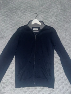 Mörkblå ziptröja från Pier One - Mörkblå ziptröja från Pier One med hel dragkedja och ståkrage. Tröjan har grått innerfoder i kragen och två fickor framtill. 
