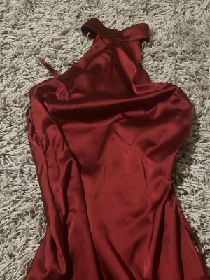 Röd kort satinklänning med halterneck - Snygg kort röd  klänning i glansigt satintyg med halterneck och öppen rygg. Klänningen har en figurnära passform och ett bälte i midjan där bak  för extra detalj. Perfekt för dig som vill sticka ut med en elegant look.