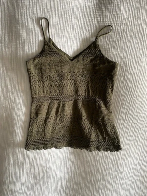 Olivgrön spetslinnetopp med smala band - Olivgrön linnetopp med smala axelband och v-ringning. Toppen är täckt av ett vackert spetsmönster och har en feminin, lätt figurnära passform. Perfekt att styla med jeans eller kjol för en trendig look.
