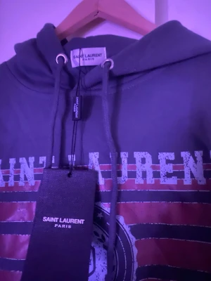 Saint Laurent hoodie med tryck - Cool hoodie från Saint Laurent Paris i mörk färg med tryckt logga och grafiskt mönster framtill. Klassisk huva med dragsko och ribbade muddar. Perfekt för dig som gillar streetstyle och exklusiva märken.