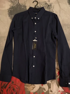 Marinblå skjorta från Polo - Snygg mörkblå skjorta från Polo Ralph Lauren med klassisk krage och vita knappar. Skjortan har lång ärm och är tillverkad i ett tunt, mjukt bomullstyg. Perfekt för dig som gillar stilrena och tidlösa plagg.