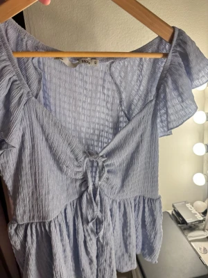 Ljusblå blus med knyt och volang - Supersöt ljusblå topp med knyt framtill och volangärmar. Tunt, krinklat tyg som ger en luftig känsla. Toppen har en feminin v-ringning och är kortare i modellen.