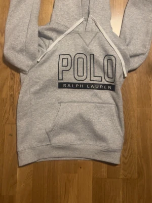 Grå hoodie från Polo Ralph Lauren - Snygg ljusgrå hoodie från Polo Ralph Lauren i mjukt bomullsmaterial. Klassisk modell med huva och ribbade muddar vid ärmslut och nederkant. Perfekt för en avslappnad och stilren look.