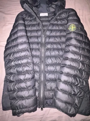  Jacka från Stone Island - Snygg svart dunjacka från Stone Island med huva och quiltad design. Jackan har dragkedja framtill, ikonisk logotyp på ärmen och är tillverkad i lätt, värmande material. Perfekt för kalla dagar och streetwear-stil. Priset är inte hugget i sten och är öppen för bud, Äkta vara ifrån Stone Island ✅