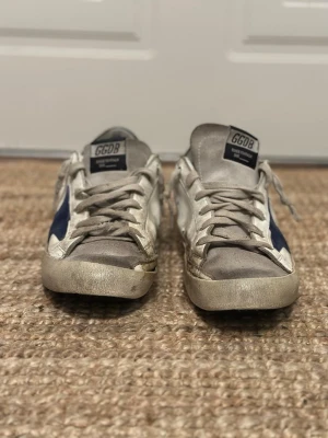 Golden Goose Superstar sneakers vit/blå - Golden Goose Superstar sneakers i vitt skinn med blå detalj på sidan och mörkblå häl. Klassisk låg modell med snörning och rund tå. Tungan har GGDB-logga och skon har en vintage-inspirerad look. Tveka inte att höra av dig med frågor o annat 😊