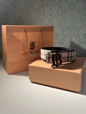Burberry bälte  med rutigt foder - Snyggt två sidade bälte från Burberry med klassiskt rutigt foder på insidan och guldfärgat BB-spänne. Bältet är gjort i läder och kommer med originalpåse och box. Perfekt accessoar för att lyfta din outfit. Pris går och diskutera.