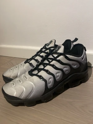 Nike Air Vapormax Plus vit/svart - Nike Air Vapormax Plus sneakers i silver och svart med tydliga svarta detaljer och snörning. Ovandelen är i syntetmaterial och har en unik, vågig design. Sulan är svart och genomskinlig med synliga luftkuddar för extra komfort. Perfekta för dig som gillar streetwear.