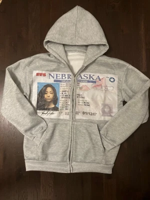 Grå hoodie med tryck - Säljer en grå zip-hoodie med stor körkort-tryck på framsidan. Hoodien har huva och dragkedja samt två fickor framtill. Perfekt för dig som gillar unika och tuffa archive plagg med statement-tryck. Materialet är bomull med ett fleece lager på insidan.