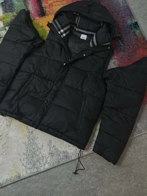 Svart pufferjacka från Burberry - Svart pufferjacka från Burberry med huva och dragkedja framtill. Jackan har klassiskt rutigt foder i huvan och logotypen 'Burberry London England' på ärmen. Två sidofickor med dragkedja och justerbar dragsko nedtill. Perfekt för kalla dagar.