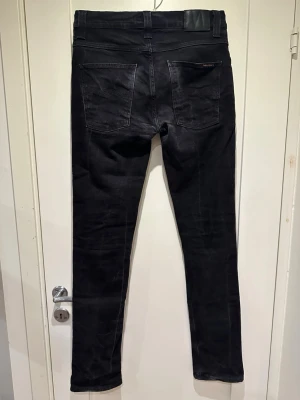 Nudie Jeans Grim Tim - Snygga svarta jeans från Nudie Jeans med klassisk femficksmodell och smal passform. Jeansen har diskreta sömmar och svart läderpatch bak i midjan. Perfekta till en avslappnad men stilren look.